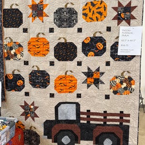 May include: A handmade Halloween-themed quilt featuring pumpkin and star patterns, along with a black truck design. The quilt measures 62.5 x 68.5 inches. The pumpkins are orange and black, with various patterns. The background is a neutral color.