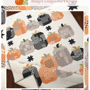 May include: A quilt featuring a repeating pattern of patchwork pumpkins in shades of orange, gray, and black, with star accents on a white background. The quilt is labeled "Hocus Pocus" and includes the website address www.thepatternbasket.com.