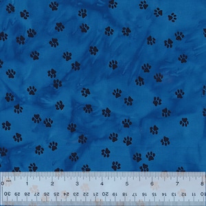 Op de afbeelding: Blauwe stof met een patroon van zwarte pootafdrukken. Onderaan de afbeelding staat een liniaal met metingen in inches en centimeters. De stof heeft een gevlekt uiterlijk.