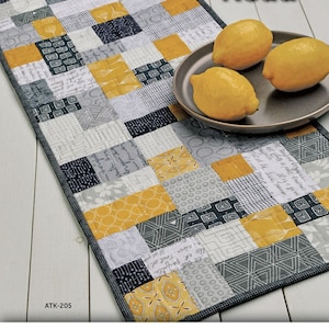 May include: A rectangular patchwork table runner with a "Mini Brick Road" design. The runner features a mix of yellow, gray, and white patterned fabric squares. Three lemons rest on a gray plate. The Atkinson Designs logo is at the bottom.