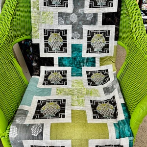 Puede incluir: Un edredón hecho a mano con un diseño de búho repetido en un patrón de patchwork. El edredón incorpora cuadrados de telas blancas, negras, grises, turquesas y verdes. El edredón está colocado sobre una silla de mimbre verde brillante.