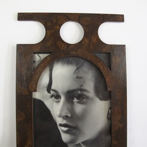 May include: A rectangular, dark brown picture frame with a decorative top featuring two circular cutouts and a horizontal extension. The frame surrounds a black and white photograph of a woman's face. The frame has a distressed, textured finish.