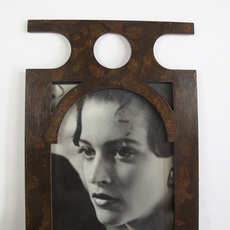 May include: A rectangular, dark brown picture frame with a decorative top featuring two circular cutouts and a horizontal extension. The frame surrounds a black and white photograph of a woman's face. The frame has a distressed, textured finish.