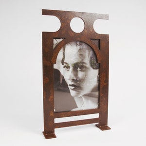 May include: A decorative, freestanding metal picture frame with a rust-colored finish. The frame features an arched opening for the photo and a geometric design at the top. The black and white photo inside shows a woman's face.