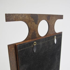 May include: A vintage-style, rectangular picture frame with a distressed, rusty brown finish. The frame features a unique top design with two circular cutouts and a rectangular top. The back is covered in black fabric, with hanging hardware.