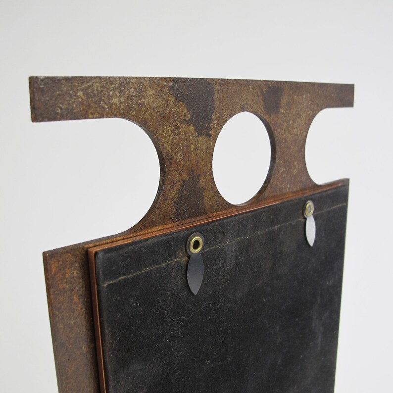May include: A vintage-style, rectangular picture frame with a distressed, rusty brown finish. The frame features a unique top design with two circular cutouts and a rectangular top. The back is covered in black fabric, with hanging hardware.