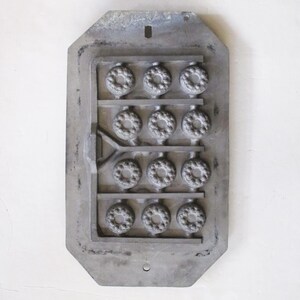 May include: A rectangular, gray metal mold with a series of circular cavities arranged in rows. Each cavity features a detailed, floral-like design. The mold has a textured surface and is likely used for casting small decorative items.