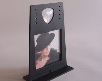 Puede incluir: Soporte negro con un pequeño marco de fotos y una gema transparente en forma de triángulo. El marco de fotos muestra a una mujer con un sombrero negro.