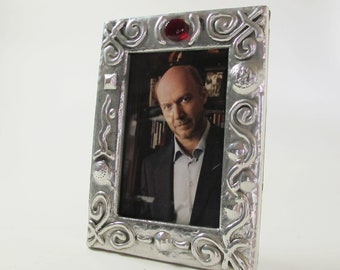 May include: A silver picture frame with an ornate design and a red glass accent. The frame is holding a photo of a man in a suit.