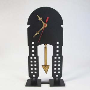 May include: Black clock with a gold pendulum and hands. The clock is shaped like a building with a black face and a white background. The clock is on a black stand.