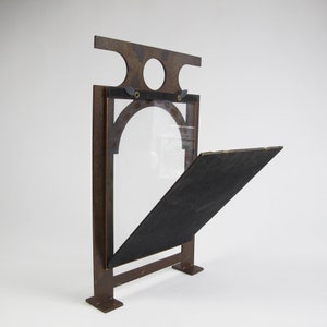 May include: A decorative, open-faced sign with a dark brown metal frame. The frame has an arched window with a clear glass pane and a hinged, angled black backing. The top of the frame has two circular cutouts.