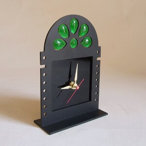 May include: A black, art deco-style desk clock with a square face and a decorative arched top. The clock features gold hands, a red second hand, and green glass accents. The clock is made of wood and sits on a rectangular base.