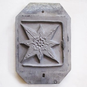 May include: A weathered, gray metal plaque with an octagonal shape. The plaque features a raised, eight-pointed star design within a rectangular frame. The star's points are adorned with decorative elements, and the center has a textured pattern.