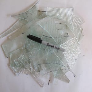 May include: A collection of clear, textured glass shards in various geometric shapes and sizes, featuring ribbed, dotted, and swirled patterns. A black Sharpie Fine Point marker is placed horizontally across the center of the glass pieces.