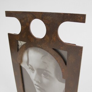 May include: A decorative photo frame with a distressed, rusty brown finish. The frame features a geometric design with circular cutouts at the top and an arched opening for the photo. The frame holds a black and white image of a woman.