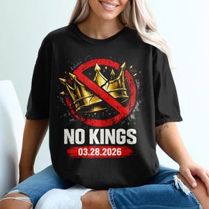 以下が含まれることがあります： 黒いTシャツ。赤い円で打ち消された金の王冠のグラフィックデザインが特徴です。「NO KINGS」の文字がグラフィックの下にあり、「03.28.2026」の日付が赤いバナーに表示されています。カジュアルウェアアイテムです。
