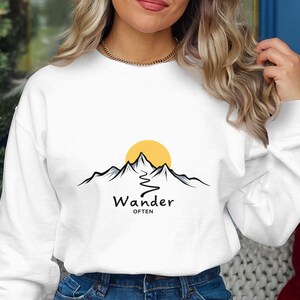 以下が含まれることがあります： 山と太陽のグラフィックと「Wander Often」の文字が黒でプリントされた白いスウェットシャツ。デザインには曲がりくねった道が含まれています。カジュアルなスタイルのスウェットシャツ。