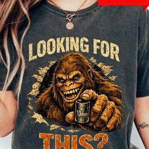 Puede incluir: Camiseta gris oscuro con un gráfico de Bigfoot sosteniendo un dado de 10 mm. El texto "LOOKING FOR THIS?" está impreso en letras naranja desgastadas. El diseño incluye una etiqueta PNG & SVG.