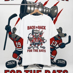 May include: White t-shirt with a hockey-themed graphic. The design features cartoon mice in hockey uniforms holding a trophy, with the text "BACK TO BACK" and "FOR THE RATS". Hockey pucks and hockey players are also in the design.