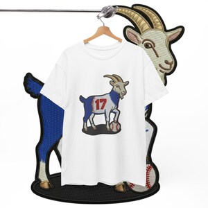 May include: White t-shirt featuring a cartoon goat with blue and red accents, wearing the number 17, and standing on a baseball. The shirt is hanging on a wooden hanger. A goat patch is behind the shirt.