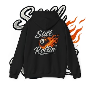 Puede incluir: Sudadera con capucha negra con el texto "Still Rollin'" y un gráfico de una bola ocho en llamas. El diseño es en blanco y naranja. La sudadera tiene capucha y bolsillo delantero.