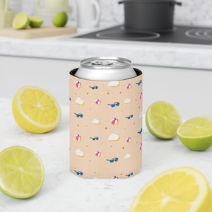 Puede incluir: Un enfriador de latas beige con un patrón repetido de gafas de sol, pelotas de playa, nubes y puntos. El enfriador sostiene una lata plateada. Limones y limas en rodajas están esparcidos alrededor del enfriador sobre una superficie blanca.