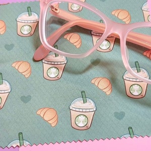 May include: A pink microfiber cleaning cloth with a green background and a repeating pattern of coffee cups and croissants. A pair of pink-rimmed glasses is lying on top of the cloth.