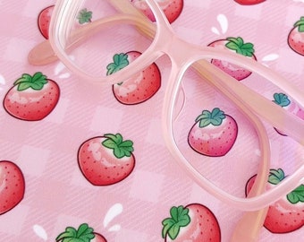 May include: A pair of light pink eyeglasses with a cat-eye frame rests on a pink strawberry-patterned cloth. The cloth has a gingham pattern with cartoon strawberries. The glasses have a subtle, translucent appearance.