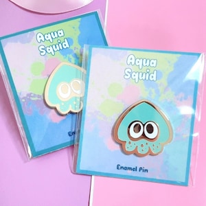 May include: Two Aqua Squid enamel pins on their packaging cards. The pins are light blue and gold with a cartoon squid design. The packaging cards have a watercolor background with the text "Aqua Squid" and "Enamel Pin".