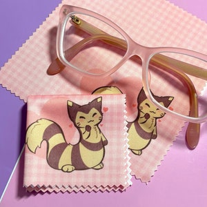 May include: Two pink microfiber cleaning cloths with a cartoon illustration of a yellow and brown cat with a pink heart on each cloth. A pair of pink cat-eye glasses are on top of the cloths.