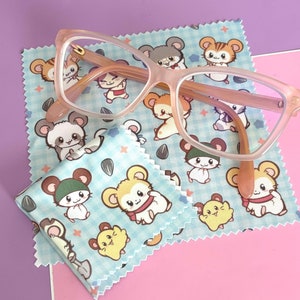 May include: Pale pink cat-eye glasses sit on two blue cleaning cloths decorated with a cartoon hamster pattern. The background is a combination of purple and pink. The cloths are made of a soft material.