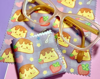 May include: A pair of pink-rimmed glasses with a brown frame sits on top of two purple microfiber cleaning cloths. The cloths have a pattern of cartoon pudding cups with smiling faces and strawberries.