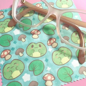 May include: A blue and green microfiber cleaning cloth with a repeating pattern of frogs, mushrooms, and lily pads. The cloth is designed to clean eyeglasses.