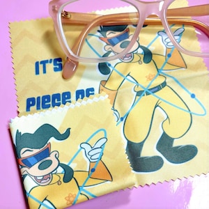 May include: Pale pink spectacles sit on a yellow cleaning cloth with a cartoon character in a yellow jumpsuit and the text "IT'S Piece of". The cloth has a zigzag pattern and a cartoon character illustration.
