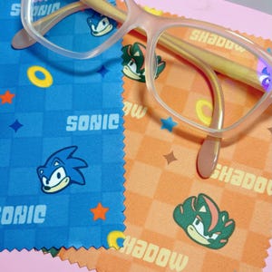 May include: Pale pink eyeglasses sit on two cloths. One is blue with the word "SONIC" and a cartoon hedgehog. The other is orange with the word "SHADOW" and a green hedgehog. The eyeglasses have clear lenses and a light pink frame.