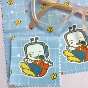 May include: Light blue microfiber cleaning cloths featuring a cartoon TV character design, red hearts, and a grid pattern. The cloths are next to a pair of pink-framed spectacles. The TV character is wearing a red jacket and holding a microphone.
