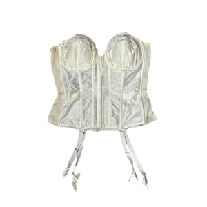 May include: Ivory-colored vintage corset with a structured design. Features a front closure, boning, and delicate lace trim along the bustline. Includes attached garter straps. The corset is made of a satin-like material.