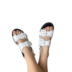 May include: White leather sandals with black soles. The sandals feature two adjustable straps with silver-colored buckles. The footwear is designed with a minimalist aesthetic, suitable for casual wear.