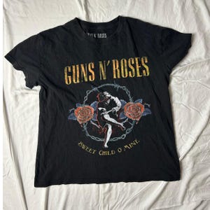 Könnte beinhalten: Schwarzes Guns N' Roses T-Shirt mit dem Bandlogo in goldener Schrift. Das Shirt zeigt eine Grafik einer Figur, Rosen und den Text "Sweet Child O' Mine". Das T-Shirt ist aus weichem Material.