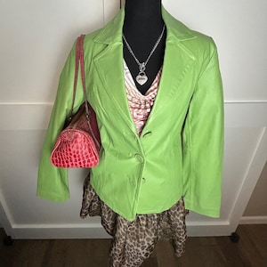 May include: A lime green leather blazer with a single-button closure, paired with a patterned top and a leopard print skirt. A red handbag with a crocodile skin texture hangs on the left side. A silver necklace with a heart pendant is visible.