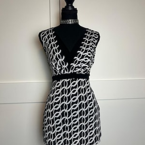 May include: A sleeveless dress featuring a black and white interlocking chain-link pattern. The garment has a deep V-neckline with solid black trim, a defined black waistband, and a straight hemline that falls above the knee on a mannequin.