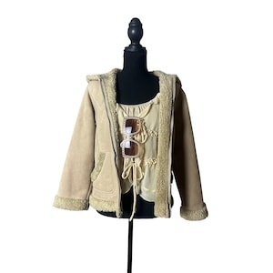 May include: A beige hooded jacket with a fuzzy interior lining and trim, worn over a light yellow sleeveless top featuring a lace-up front detail. A pair of brown-tinted sunglasses with decorative rhinestone frames rests on the top.