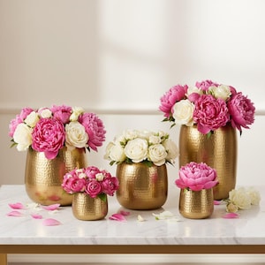 Könnte beinhalten: Vier gehämmerten goldfarbenen Metallvasen in verschiedenen Größen, jeweils gefüllt mit Arrangements aus rosa und weißen Pfingstrosen und Rosen. Die Vasen haben eine abgerundete Form und eine strukturierte Oberfläche mit verstreuten Blütenblättern.