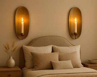 May include: Two oval-shaped brass wall sconces with lit taper candles mounted on a wall above a bed. The sconces feature a smooth, metallic finish and a central candle holder. The light from the candles casts a warm glow on the wall.