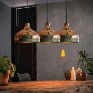 May include: Three copper-coloured pendant lights with a green patina hang above a wooden table. The lights are suspended from black cords. The logo "ACHGALO" is visible in the bottom right corner. The room has a neutral colour scheme.