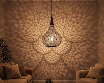 May include: A decorative pendant light fixture with a teardrop shape, featuring intricate cut-out patterns. The light casts a patterned design on the surrounding walls. The fixture hangs from a chain and illuminates a living room setting with neutral-toned furniture.