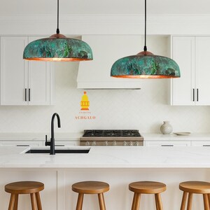 May include: Two dome-shaped pendant lights with a green and copper finish hang above a kitchen island. The lights are suspended by black cords. The kitchen features white cabinets, a black faucet, and wooden stools.