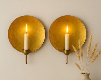 May include: Two gold-colored wall sconces with lit white candles. Each sconce has a round, textured plate and a candle holder. A vase with wheat stalks is in the lower right corner. The sconces are mounted on a light-colored wall.