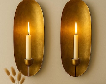 May include: Two gold-colored wall sconces with lit candles. Each sconce has an oval shape and a small candle holder. The candles are white and the flames are bright. A small white vase with dried flowers and a small bowl are in the foreground.