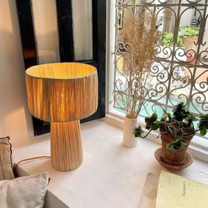 Peut inclure: Une lampe en osier naturel avec un abat-jour rond de couleur brun clair et une base cylindrique haute. La lampe est allumée et diffuse une lumière chaude. La lampe est posée sur une surface blanche à côté d'une fenêtre donnant sur une cour.